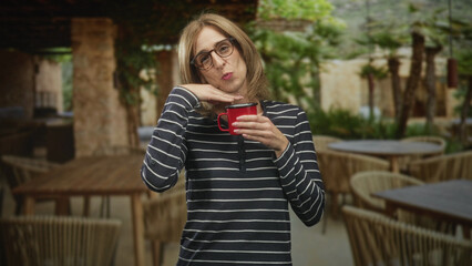 Fototapeta premium Woman holding red mug with hand under chin gesture, wearing glasses and striped shirt in a building cafe patio; playful delight.