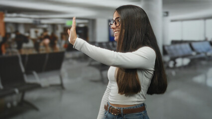 Woman waves hand in airport terminal, smiling and looking sideways, wearing glasses, white long sleeve top, jeans and belt, long hair visible  farewell joy. © Krakenimages.com