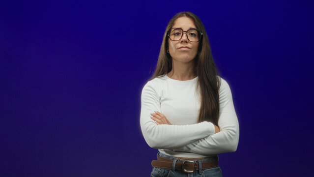 Woman with glasses, arms crossed, long hair, wearing white top, jeans and brown belt against a deep purple studio backdrop; quiet determination.