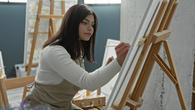 Young latin woman painter wearing apron leans forward and paints a canvas with a brush on a wooden easel in a studio; creativity concentration.