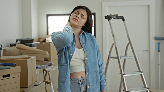 Woman rubbing neck and wincing amid stacked cardboard boxes, ladder and tools during moving in a building; relocation fatigue.