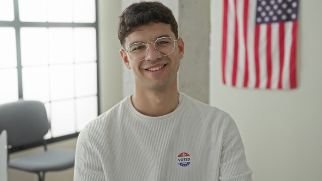 Man smiling and holding smartphone with an 'i voted' sticker visible inside a civic building with american flag on wall; civic pride.