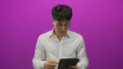 Man smiling and laughing while holding a tablet with both hands and tapping the screen in a purple studio wearing round glasses and a white shirt  joy curiosity. © Krakenimages.com