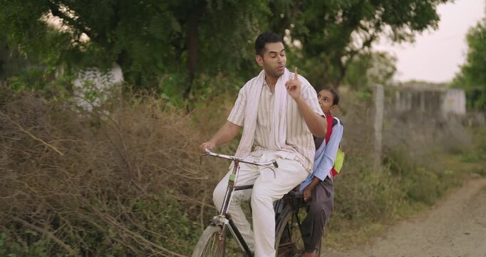 Indian happy young adult dad talk speak drop school to teen girl child rural area place road path way. Smiling female wear uniform with bag sitting on cycle back seat enjoy ride have fun joy desi life