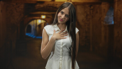 Fototapeta premium Young woman in white dress places hand on chest with soft smile and long hair visible in an arched underground stone corridor; serenity reflection.