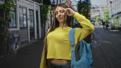 Fototapeta premium Teenage girl wearing a yellow sweater and blue backpack smiles with braces and points finger on city street; youthful confidence.