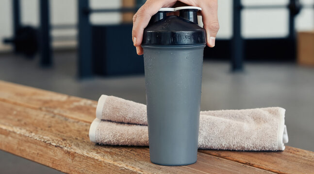 Gray shaker bottle sits on wooden bench next to folded towels. Hand holds bottle lid, preparing post workout drink. Gym interior background, fitness lifestyle.