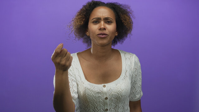 Woman beckoning with hand and furrowed brow in purple studio wearing white knit top; confusion doubt.
