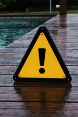 Fototapeta premium Yellow triangular warning sign with exclamation mark placed on wet wooden pier near water. Sign alerts for slippery surface on rainy day. Safety caution in public park area.