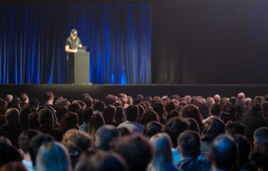 Crowd watching presenter on stage during conference, blue curtains, dim lighting, focused atmosphere, professional event, public speaking and tech presentation. © Anton Gvozdikov
