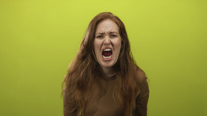 Fototapeta premium Woman with long red hair and open mouth yelling intensely in studio against a lime green background; anger frustration.