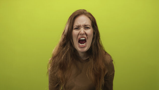 Woman with long red hair and open mouth yelling intensely in studio against a lime green background; anger frustration.