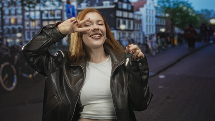 Fototapeta premium Redhead woman flashing peace sign and holding keys on a busy european street, smiling and laughing in a leather jacket; carefree joy.