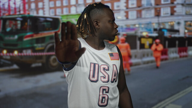 Man wearing usa jersey with hand raised palm out on street by a construction site, facing forward and pointing; defiance.