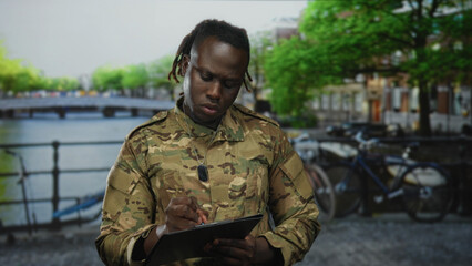 Obraz premium Man soldier in camouflage uniform writing on clipboard with pen on an amsterdam street by a canal bridge outdoors; duty focus discipline.