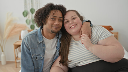 Fototapeta premium Plus size woman sits on bed with arm around man, smiling toward camera, holding his hand and showing ring in a bright bedroom building; love together.