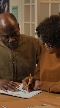 Medium vertical shot of devoted African American father homeschooling teenage son in grammar, explaining difficult spelling or sentence, boy understanding and smiling with joy