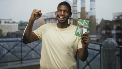 Fototapeta premium Man holding green gift with white bow and raised fist at building railing, smiling broadly; celebration victory.
