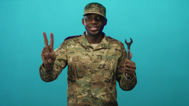 Soldier man in camouflage uniform smiles holding wrench and displays finger counts in studio; pride duty.