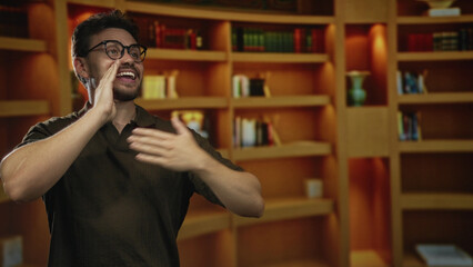 Man cupping hand to mouth calling out toward bookshelves in a warm library interior while smiling and gesturing upward  excitement learning. © Krakenimages.com