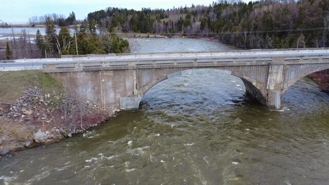Low-altitude drone shot beneath a deteriorating historic road bridge strengthened with steel supports during spring flooding in eastern Quebec, Canada.