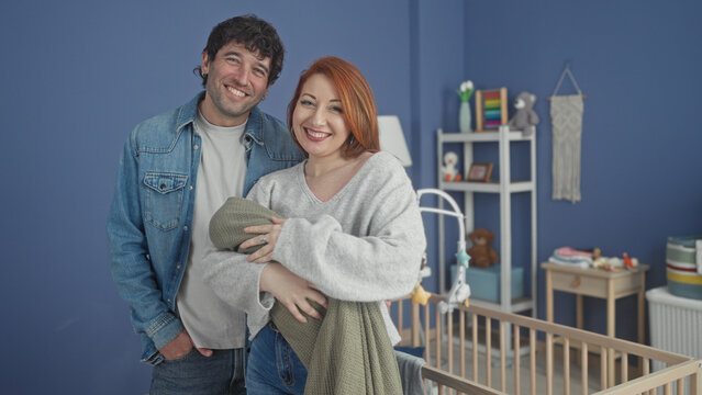 Couple cradling a swaddled baby in a home nursery with wooden crib, mobile and shelves; new parent joy.