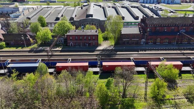 Aerial view of a railway station with freight cars and shipping containers on tracks, historic red brick industrial buildings with sawtooth roofs, green trees, sunny day with bright natural lighting, 