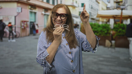 Fototapeta premium Elderly woman smiling, holding glasses and pointing finger up on a pedestrian street by cafe umbrellas and planters near pastel buildings; joyful discovery.