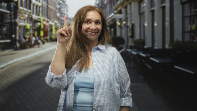 Senior woman pointing index finger and smiling on a narrow cafe street wearing light blue shirt and casual attire; friendly reminder.
