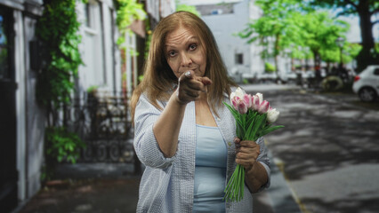 Fototapeta premium Senior hispanic woman holding pink tulips points finger directly at camera on residential street lined with trees and parked cars; assertion determination.