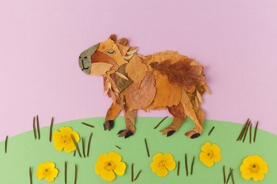 Capybara made from dried autumn leaves standing on green paper grass with yellow flowers on pink background. Nature leaf collage craft. Spring and autumn season concept. Flat lay with copy space,
