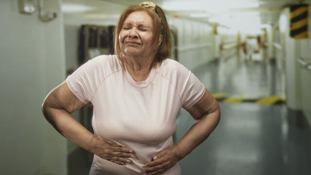 Senior hispanic woman with hands on hips wearing pink tshirt in building corridor resembling ship passage, looking resolute and steady; determination resilience.