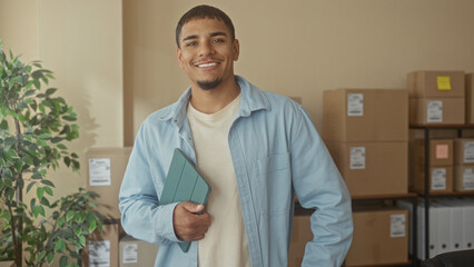 Fototapeta premium Man holding tablet to chest, smiling and standing by stacked delivery boxes and plant in a building; entrepreneurial confidence growth.
