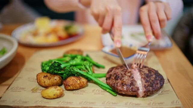 A cooked burger patty is being cut on a serving board alongside vegetables such as broccoli and small roasted potatoes
