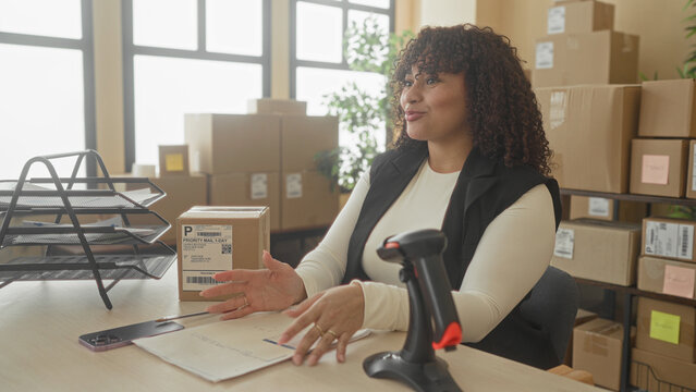 Woman claps hands beside a cardboard package and barcode label on a desk in building, smiling and gesturing near a barcode scanner and stacked shipping boxes; small business pride.