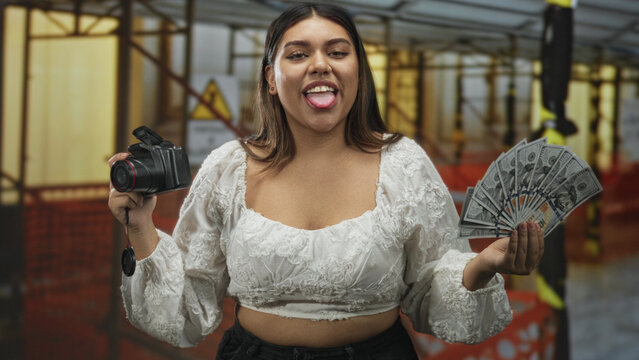 Woman holding dslr camera and fanning us dollars, showing midriff in a building; confidence monetization.