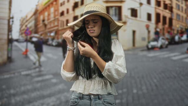 Woman holding hair between fingers and inspecting split ends, frowning in sun hat on cobblestone street with blurred buildings; frustration.