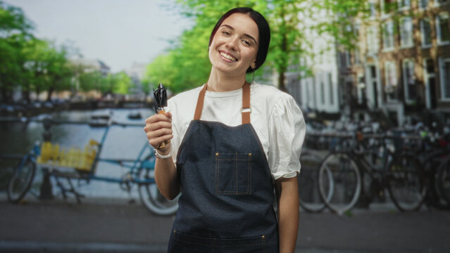 Woman gardener wearing apron holding pruning shears in hand and smiling while posing on a street by canal and bicycles; nurture joy.