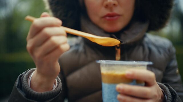 A woman wearing a dark jacket holds a small plastic cup filled with chia seed pudding while using a wooden spoon to scoop from the top