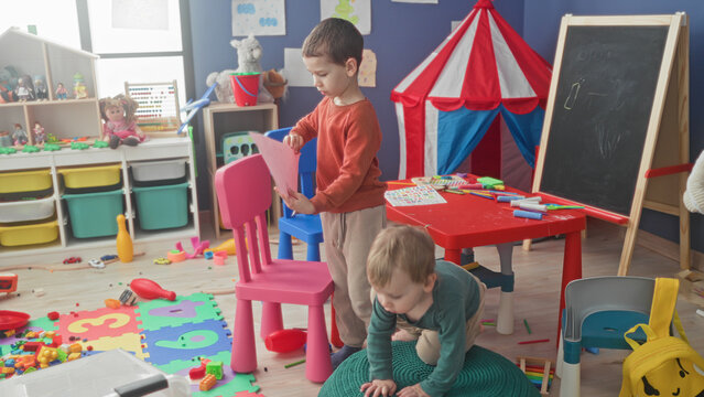 Boy holds pink paper with tongue out while younger brother sits by scattered toys inside building; childhood curiosity.