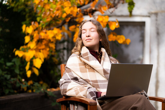 A young woman is resting her eyes after working on a laptop on a cozy autumn terrace in the daylight. The front view portrait.