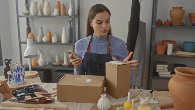 Woman wearing apron holds phone with one hand while touching cardboard box on workbench in artisan studio; concentration.