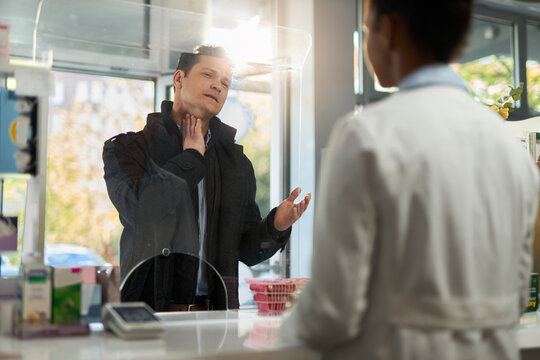 Displeased man holding his throat in pain while buying medicine at checkout in a pharmacy. The view is through glass