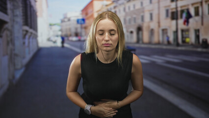 Fototapeta premium Woman clutching stomach, eyes closed, stomachache on street with blurred city buildings; nausea urgency.