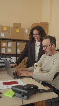 Man holding dog and smiling at laptop amid stacked cardboard boxes and parcels in a small business building; warmth teamwork.
