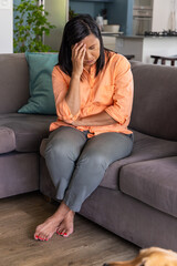 Obraz premium Mature woman sitting on gray sofa holding forehead, barefoot showing red toenails on wood floor