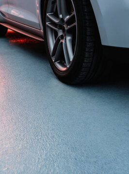 Close up of modern car wheel and tire on textured epoxy garage floor. Metallic alloy rim shows fine detail. Red underglow light reflects on clean industrial surface.