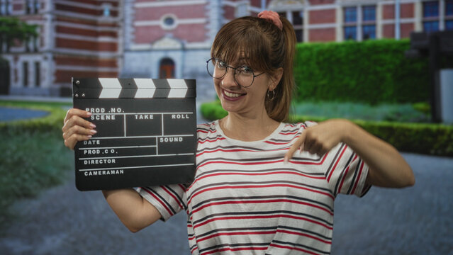 Woman holding clapperboard and pointing finger to clapperboard in front of historic building; filmmaking joy.