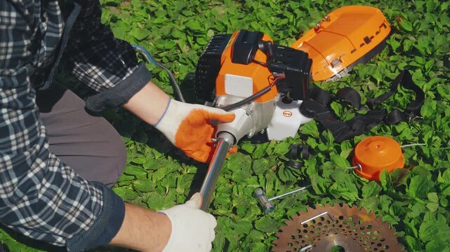 Individual in gloves works on assembling a garden tool or trimmer with a metal blade and engine on green grass, focusing on the mechanical connection and preparation for use in outdoor tasks