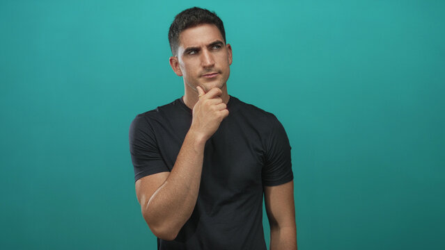 Man with hand on chin in studio with teal backdrop, wearing black tshirt and looking to the side; thoughtful reflection.